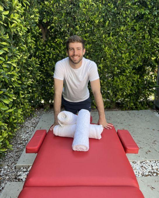 Massage therapist standing behind a treatment table with neatly rolled white towels outdoors